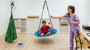 Παιδί κάθεται σε αιωρούμενη πλατφόρμα κατά τη διάρκεια άσκησης αισθητηριακής ολοκλήρωσης με τη βοήθεια θεραπεύτριας σε θεραπευτική αίθουσα.