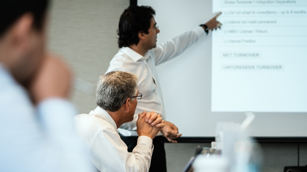 Ένας άνδρας κάνει παρουσίαση μπροστά σε προβολέα σε επαγγελματικό περιβάλλον, ενώ άλλοι παρακολουθούν.