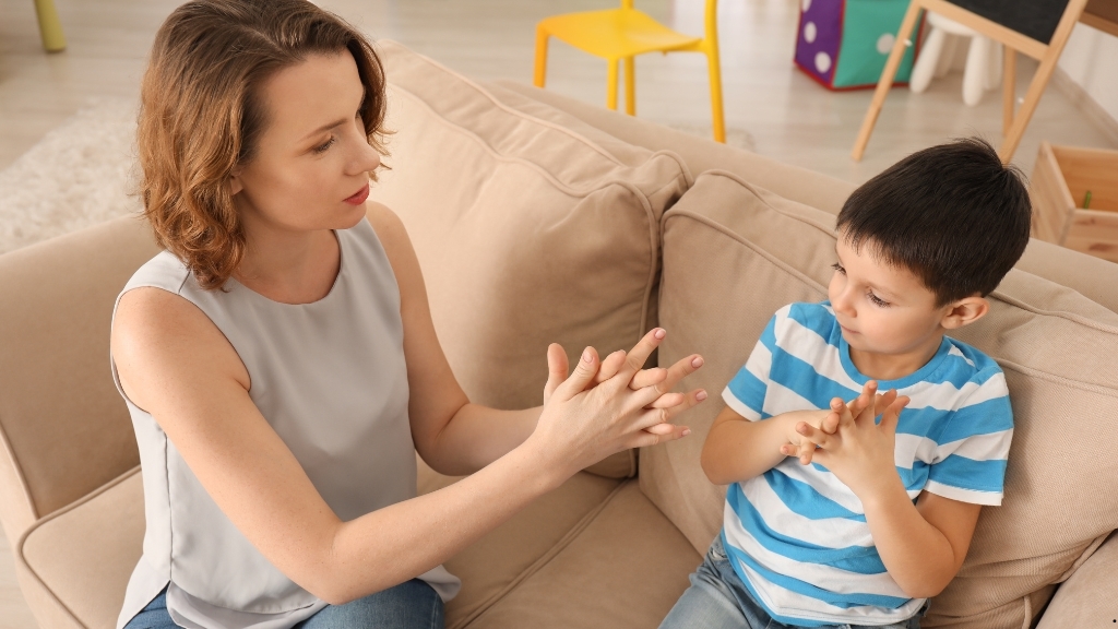 Παιδί και λογοθεραπεύτρια επικοινωνούν με νοήματα, καθισμένοι σε καναπέ.