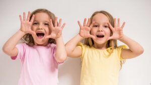 Δύο μικρά κορίτσια χαμογελούν και παίζουν, σηκώνοντας τα χέρια τους δίπλα από το πρόσωπό τους.
