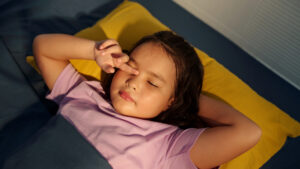 Παιδί ξαπλωμένο σε κρεβάτι, με ροζ μπλούζα, ξυπνάει και τρίβει τα μάτια του — ενδεικτικό στιγμιότυπο παιδικής ρουτίνας ύπνου.