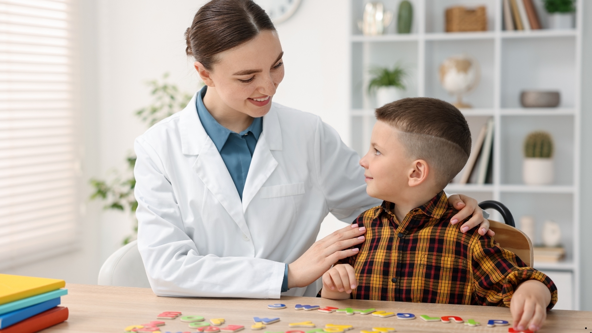 Λογοθεραπεύτρια χαμογελά σε παιδί καθώς δουλεύουν μαζί με γράμματα στο τραπέζι.