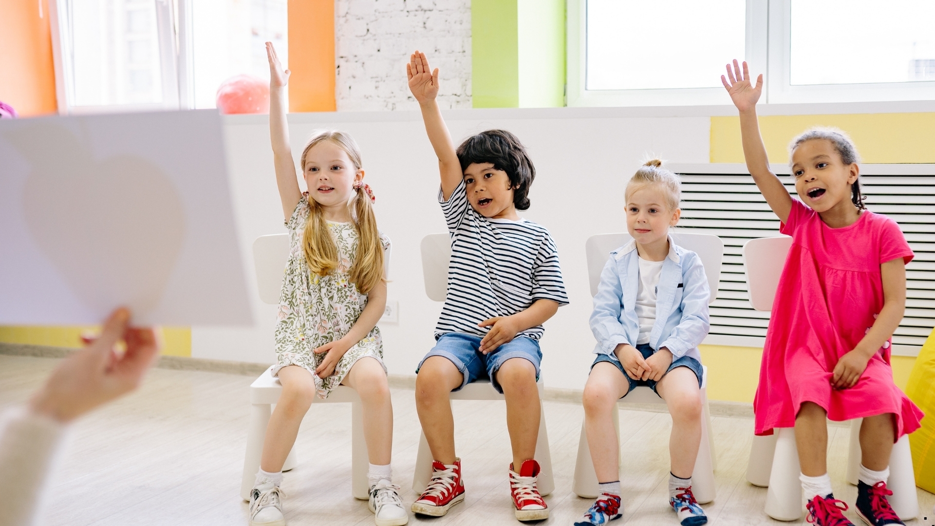 Παιδιά καθισμένα σε καρέκλες σε τάξη, με σηκωμένα χέρια, συμμετέχουν σε δραστηριότητα.